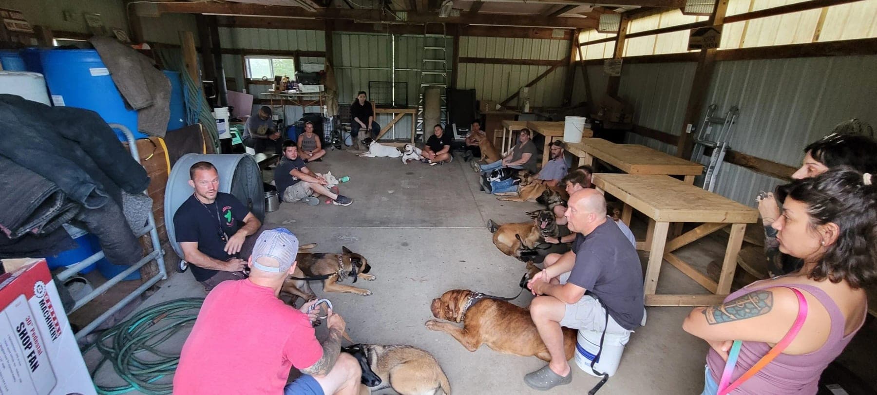 Handlers sitting in a circle on the barn floor with their dogs lying beside them